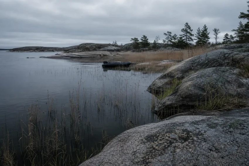 Gryning över en skärgårdsstrand med mörkt vatten, granitklippor och en svart gummibåt uppdragen bland vassen