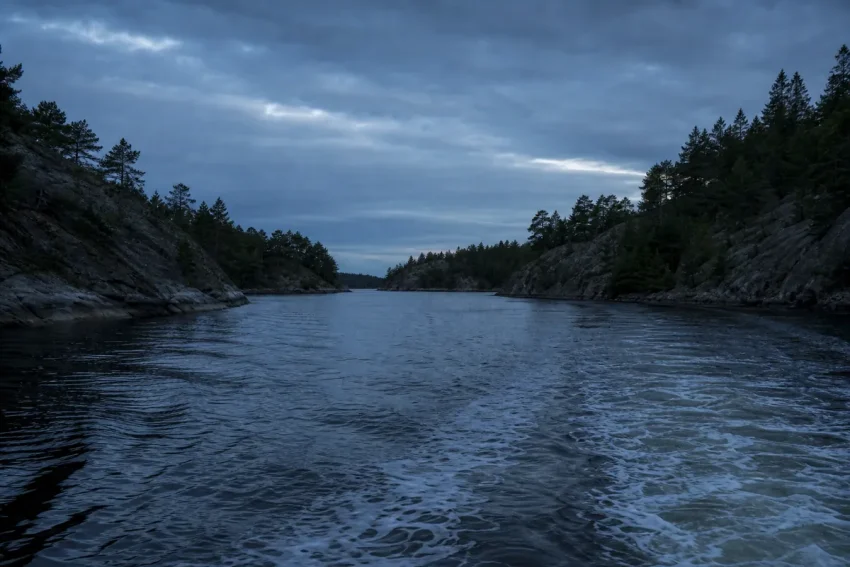 Trångt skärgårdssund i skymning sett från vattenytan med mörka klippstränder på båda sidor